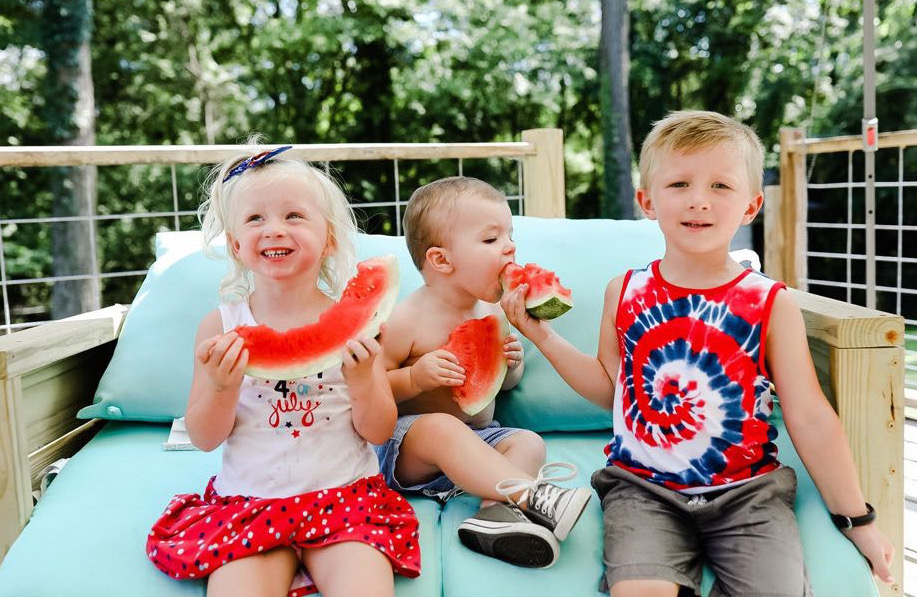 Young Rainer Lewis (age 1, in the middle) was more interested in a slice of watermelon than posing for photos on Saturday at a Fourth of July cookout celebration. Shown in the photo are (L-R) Lily Kate Mack, age 3, Rainer and Sawyer Mack, age 4, who all got plenty of watermelon, according to Ali Mercer Lewis, Rainer’s mother. 